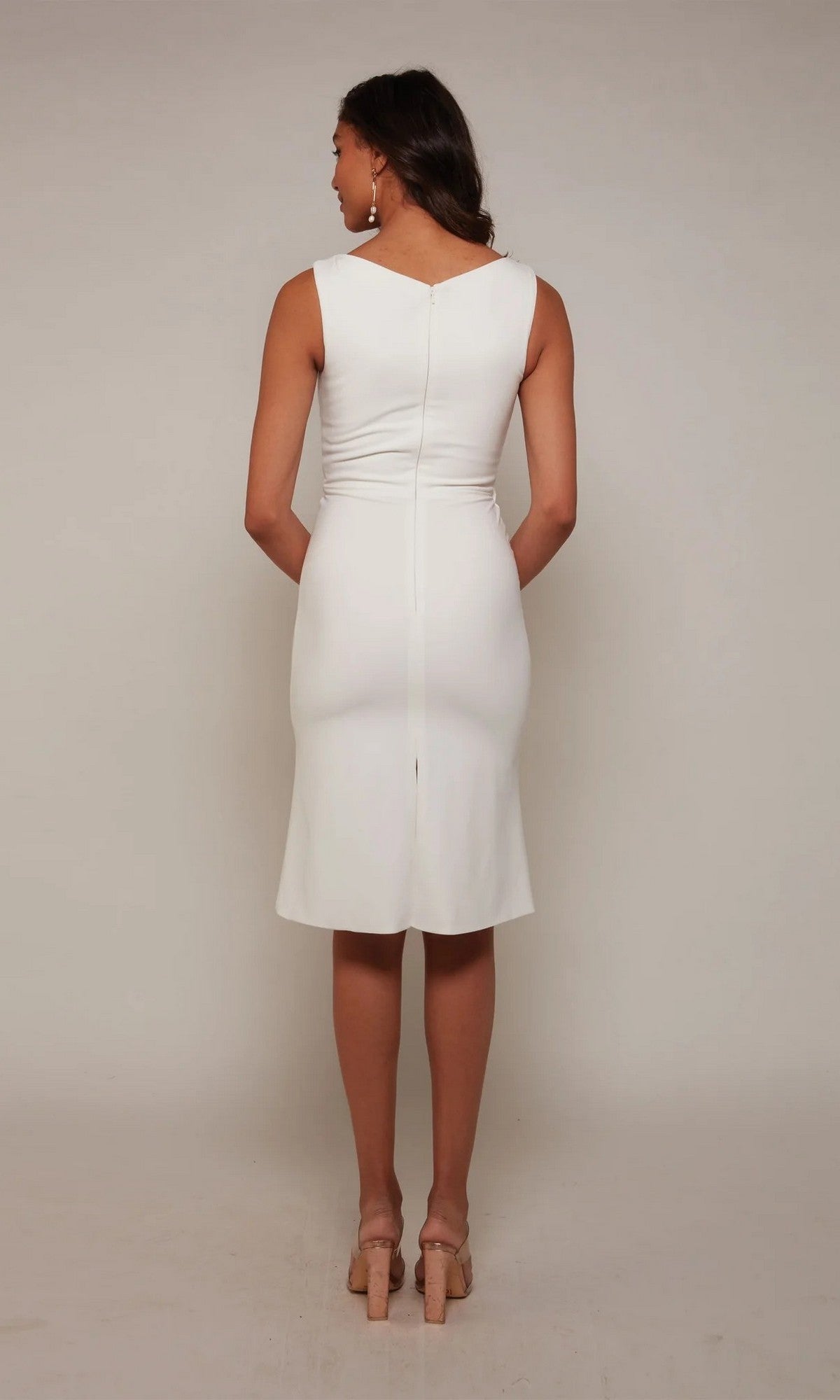 A woman faces away, wearing the Alyce 70037 knee-length white party dress with nude heels, set against a plain light background.